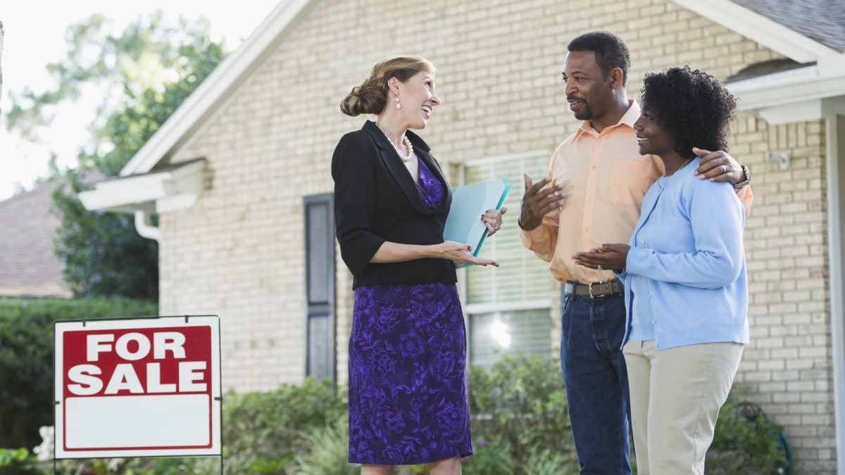 A real estate agent standing in front of a house with a For Sale sign in the yard, talking with a couple.