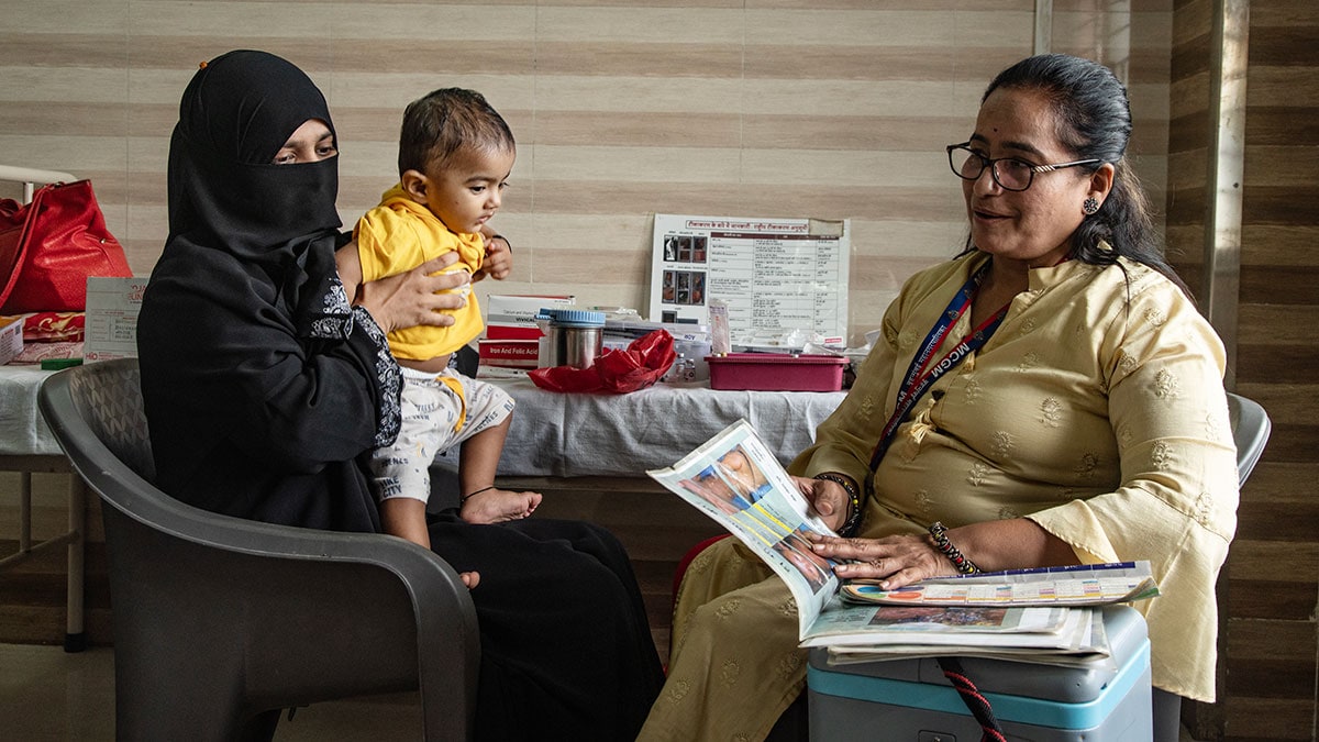A woman holds a baby while sitting across from a woman looking at a booklet.