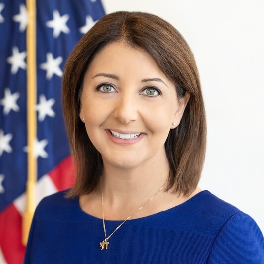 CDC Director Mandy Cohen is smiling in front of the American flag. She is wearing a blue blouse and a gold necklace.