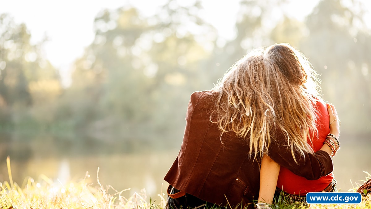 Two girls sitting on grass, facing away, hugging
