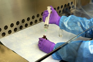 A close-up photo of the arms and hands of a worker. The hands are covered in thick purple gloves. They hold a syringe on top of a small, drug vial. The worker is also wearing a blue gown. The scene takes place on a stainless bench which has a paper cloth spread out under the vial. There is a protective glass screen between where you assume the worker%26rsquo;s face to be (cropped out of the frame) and gloved hands.