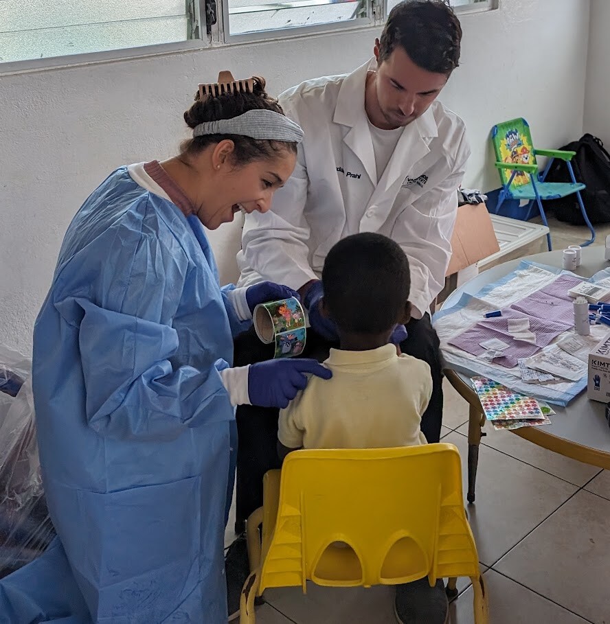 one female adult wearing gloves and a disposable robe, one adult male wearing a white lab coat and gloves, and a child with his back to the camera while he receives medical attention.