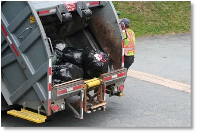 The image shows the back of a refuse truck. There are black trash bags in the back of the truck. There is a refuse worker wearing a high-visibility vest. The worker is riding on the riding step.