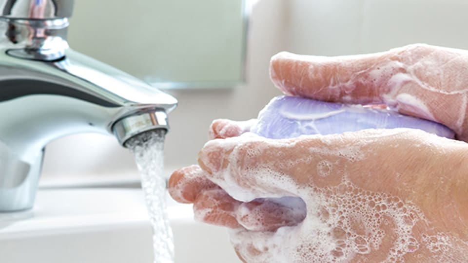 Close-up of white hands lathering with a bar of soap next to running water of a bathroom sink faucet.