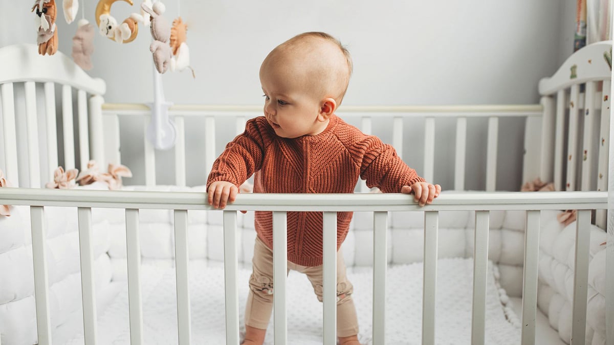 Beautiful little girl in a crib in a cozy light room in a sweater of fashionable shades. learns to stand at the support.