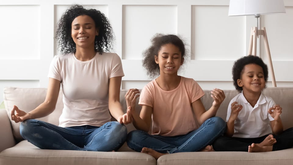Young black mother sitting cross-legged on a couch with her two small children sitting similarly next to her in meditation.