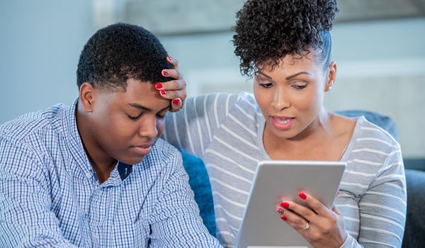 Mother feeling her teen son's forehand for a fever, while checking symptoms on a tablet