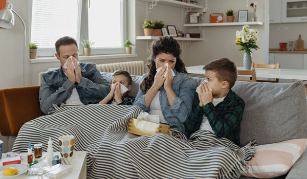 Mother and father with their two young sons, blowing their noses while sitting on the sofa under a blanket