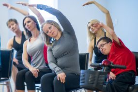 A group of people are stretching in wheelchairs inside a fitness center. They are concentrating on stretching and looking focused.