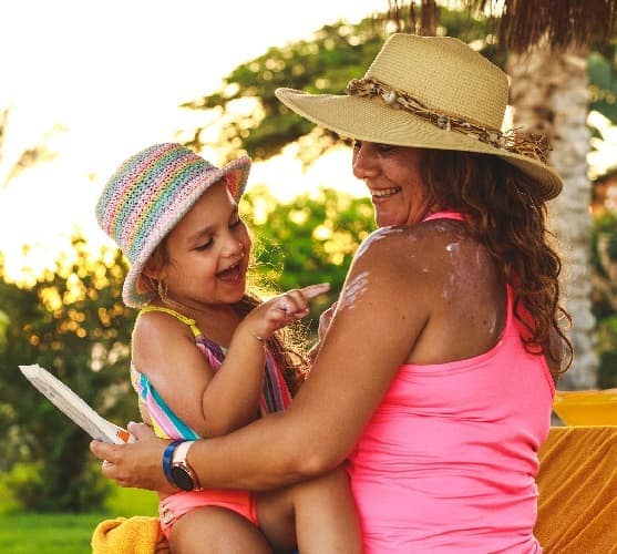 Foto de una madre y su hija en trajes de baño y sombreros y aplicándose protector solar