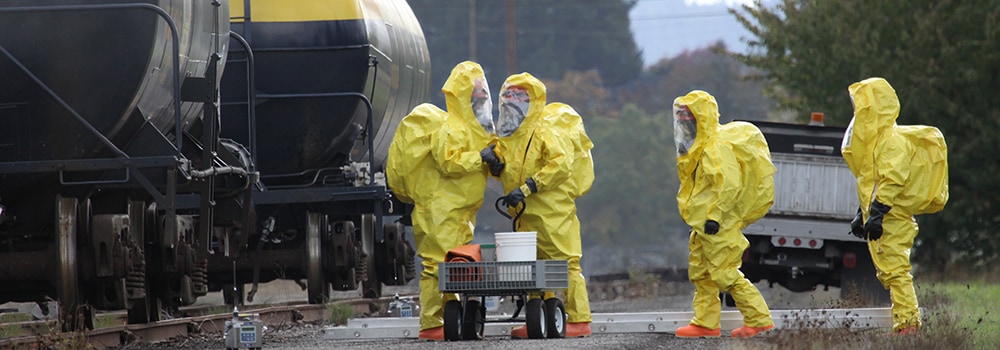 A hazmat team in yellow full-body protective gear discusses a chemical disaster.