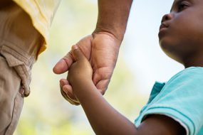 A close-up photo of a young Black child, age 3 or 4, holding the hand of an adult and looking up at them.