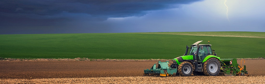 A tractor operator plows a field with lightning and dark skies in the distance