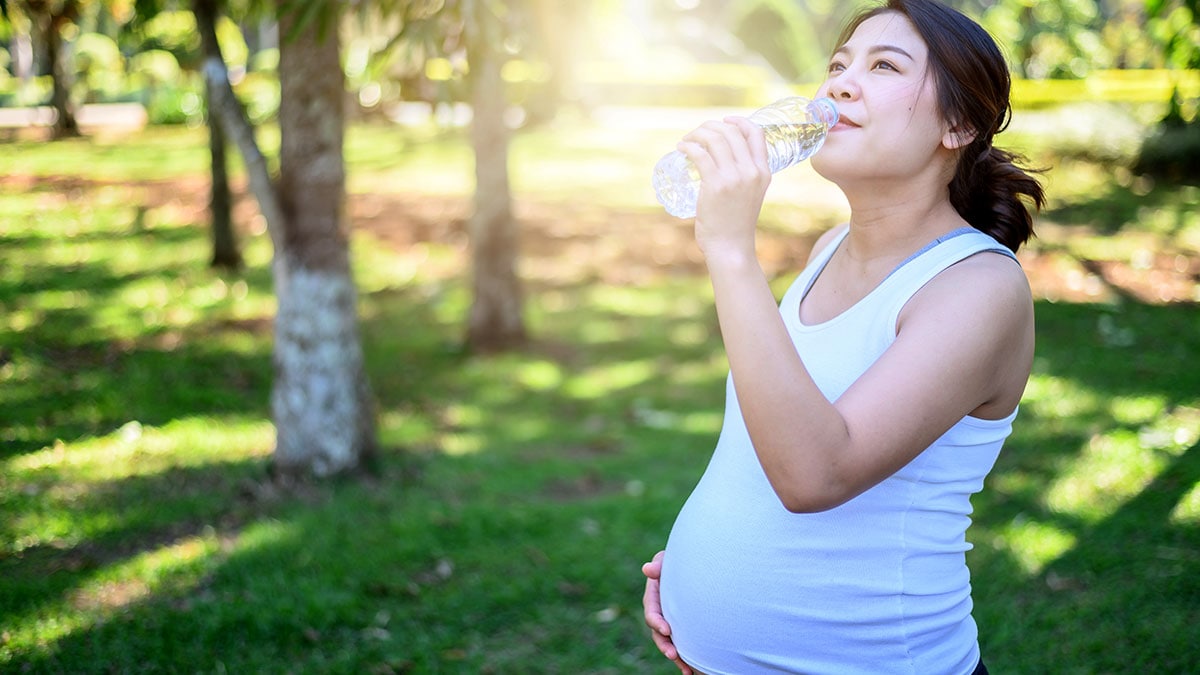 Pregnant woman is drinking water while outside on a sunny day.