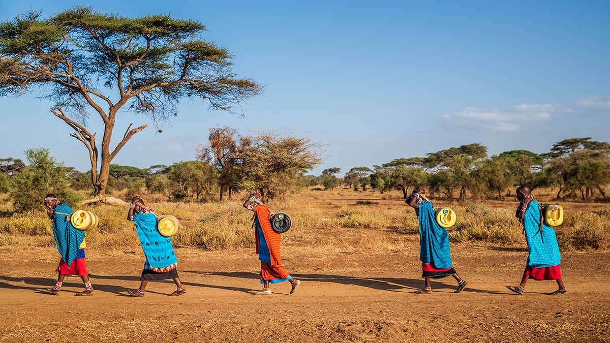 Five people wearing colorful patterned clothing walk barefoot across a dirt road. Each is holding a large jug that is slung onto their back.