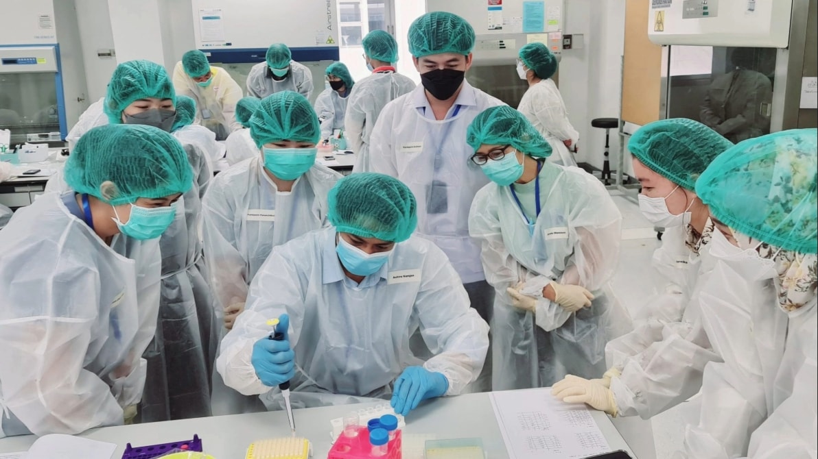 A group of people in personal protective equipment huddle around a table to watch one person who is holding a pipette over a desk.