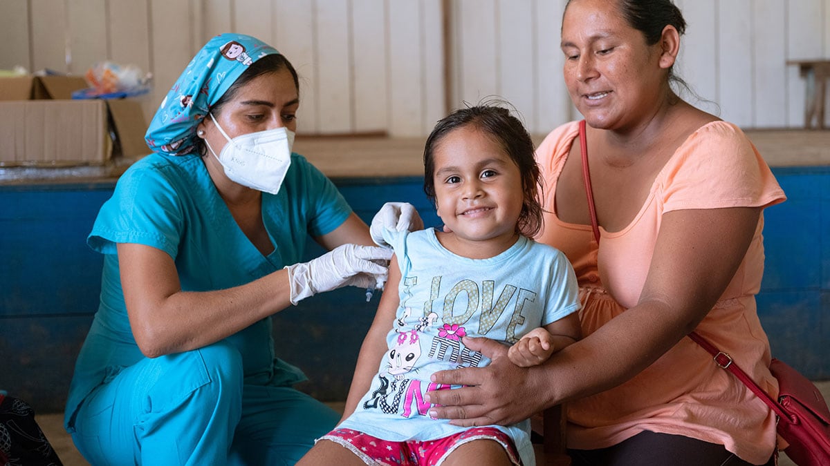A health worker vaccinates a smiling child while her mother watches.