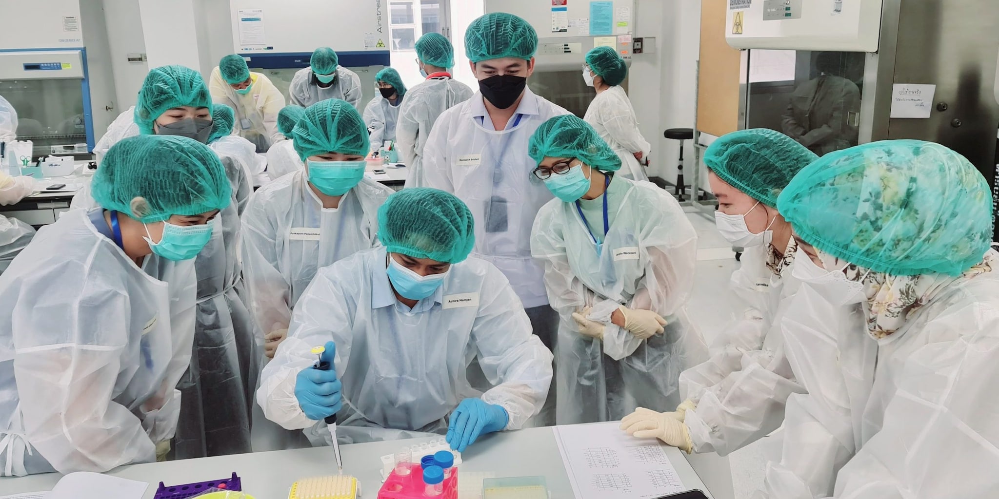Photo of 10 laboratorians watching another laboratorian hold a device used to add and remove samples from test tubes.