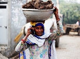 A petite woman speaks on the phone while transporting materials in a large container balanced on her head.