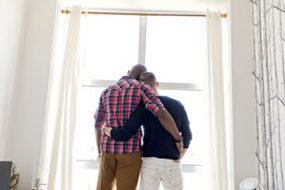 Rear view of gay men standing arms around while looking through window at home