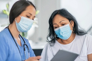 Patient with surgical mask looks at a tablet with a woman wearing gloves and a surgical mask