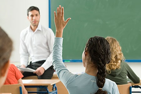 female student raises her hand to ask a question of the teacher, sitting front of class