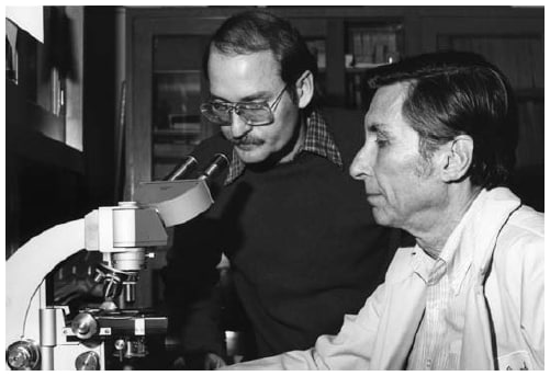 The figure is a photo of Dr. Joseph E. McDade and Dr. Charles C. Shepherd working with a microscope in a CDC lab.