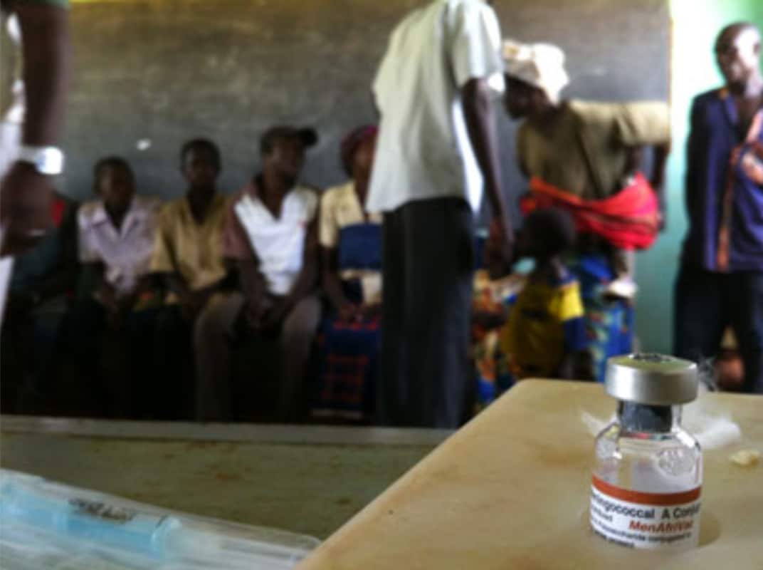 Close-up view of the MenAfriVac vaccine in a transparent bottle with a silvery lid/stopper. In the background, seven African men and women are shown seated waiting to receive the vaccine..