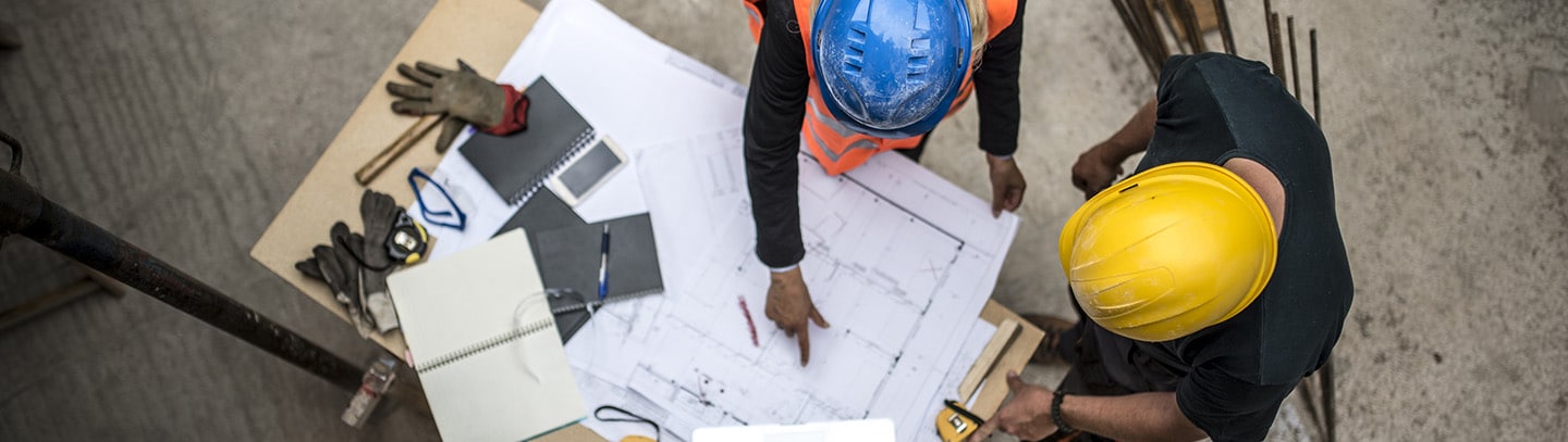 Overhead view of two construction workers in hard hats analyzing blueprints