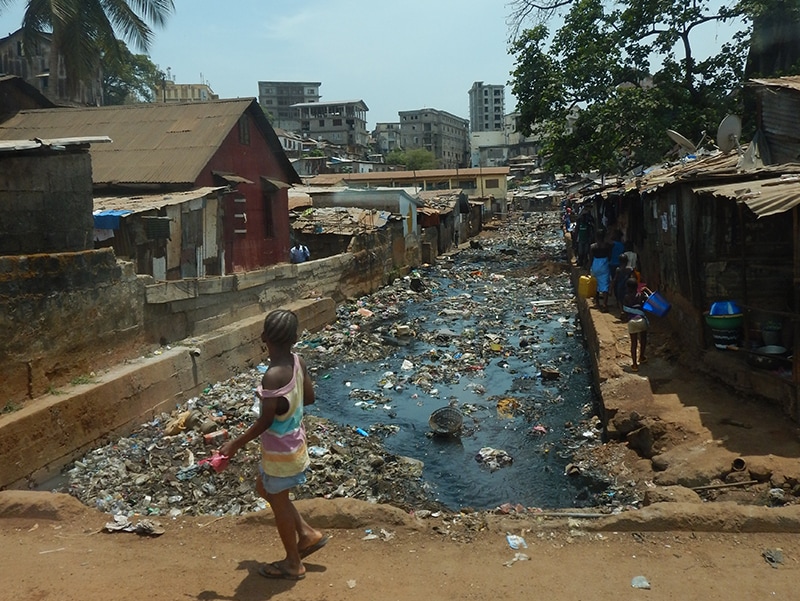 A young girl looks at a waterway that runs through a densely populated neighborhood. The waterway contains a lot of debris.