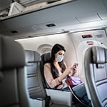 Young woman sitting using phone on the aircraft seat wearing face mask