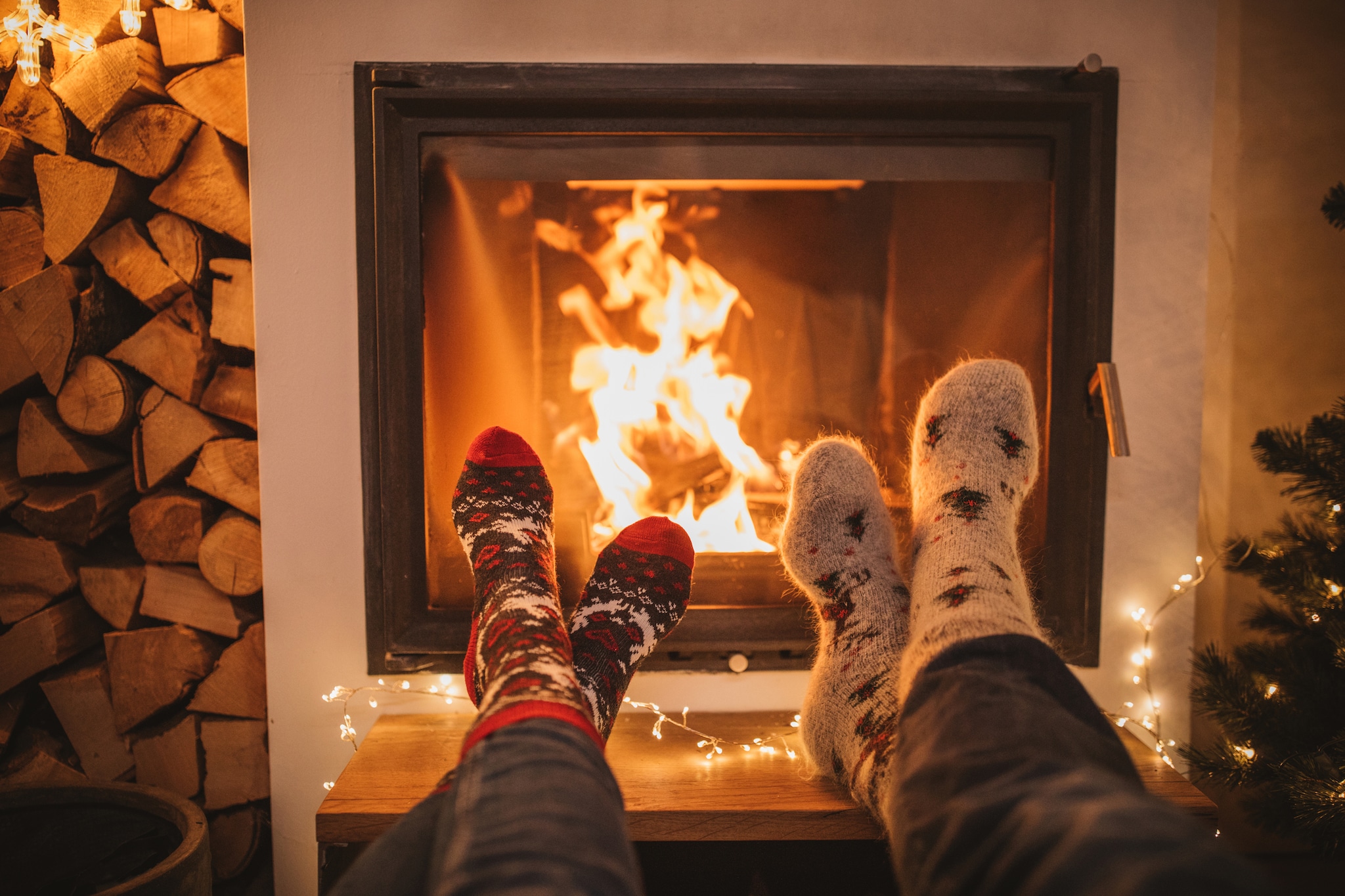 Two sets of legs with ankles crossed, wearing blue jeans and holiday print socks are stretched out on a coffee table in front of a lit fireplace.