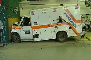 This picture is set inside an industrial looking space with concrete floor and wires and equipment attached to the walls. In the background you can see a pegboard wall with tool neatly arranged on it. In the foreground is an ambulance which has crashed head-first into a wall at the spot where there is a reddish brown plate just a little bit wider and taller than the grill of the ambulance. 