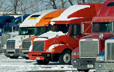 This photo was taken in the winter. Eight semi-trailers are parked in a row. You see their grills and cabs but not what they are hauling. They are different makes, models, and colors. One red semi in the center of the photo is covered with a file layer of snow. A blue semi has icicles hanging from its grill. The ground in front of them is covered with ice and snow. The trees in the background are barren and the sky is bright blue.