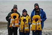 The three crew members of the setnet skiff Paul Revere posing on the shore of Bristol Bay, Alaska with the inflatable personal flotation devices that saved their lives.