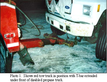 photo shows red tow truck in position with T-bar extended under front of disabled propane truck