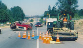 highway accident scene showing overturned truck, tractor on flatbed truck, and workzone orange cones.
