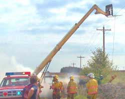 Photo 1 - View of firefighters at the scene, showing the forklift near the powerlines and the pump suspended by a wire rope cable.