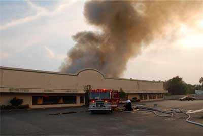 Single fire truck in front of store, with light colored smoke coming from the roof top of the building.