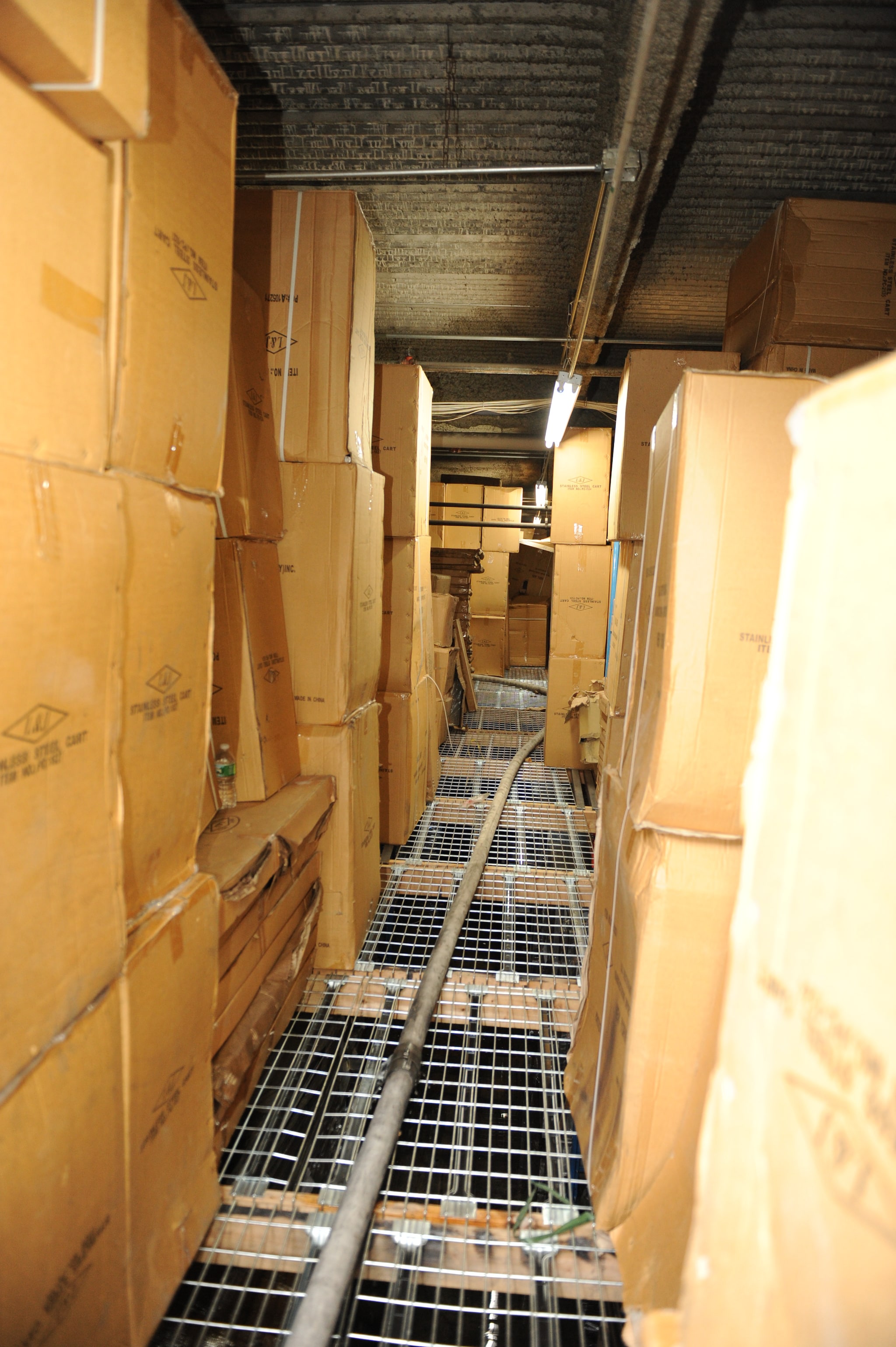 The charged attack hoseline stretched down a narrow aisle of the catwalk storage area. Large cardboard boxes are stacked from floor to ceiling on either side of the aisle.