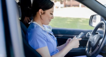 Woman in medical scrubs sits in the drivers seat of a parked car, looking at her phone. She is a White woman with dark hair.