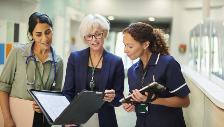 Two female healthcare workers and a older female healthcare executive gather around a discuss papers in a padfolio while standing in a hospital corridor.