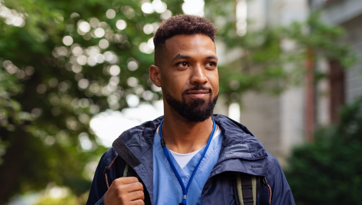 Portrait of an African American man with a  beard in medical scrubs outdoors, wearing a jacket and backpack looking hopeful.