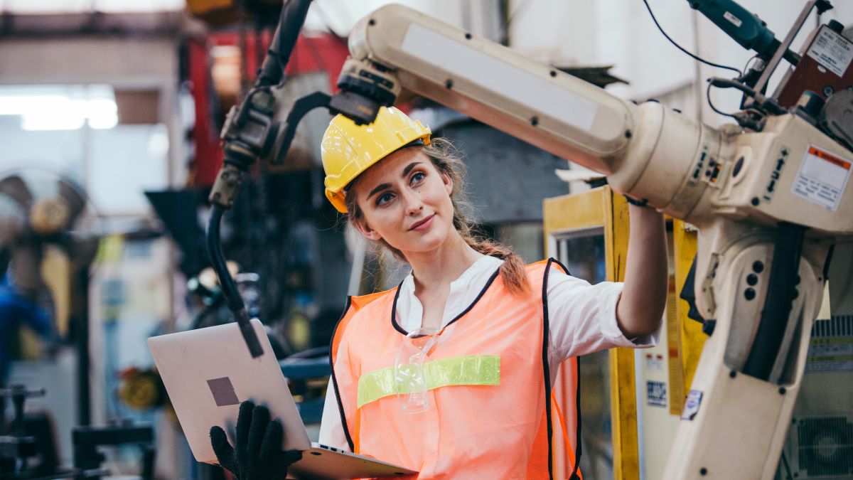 Woman wearing a hard hat and safety vest working next to a robotic arm.
