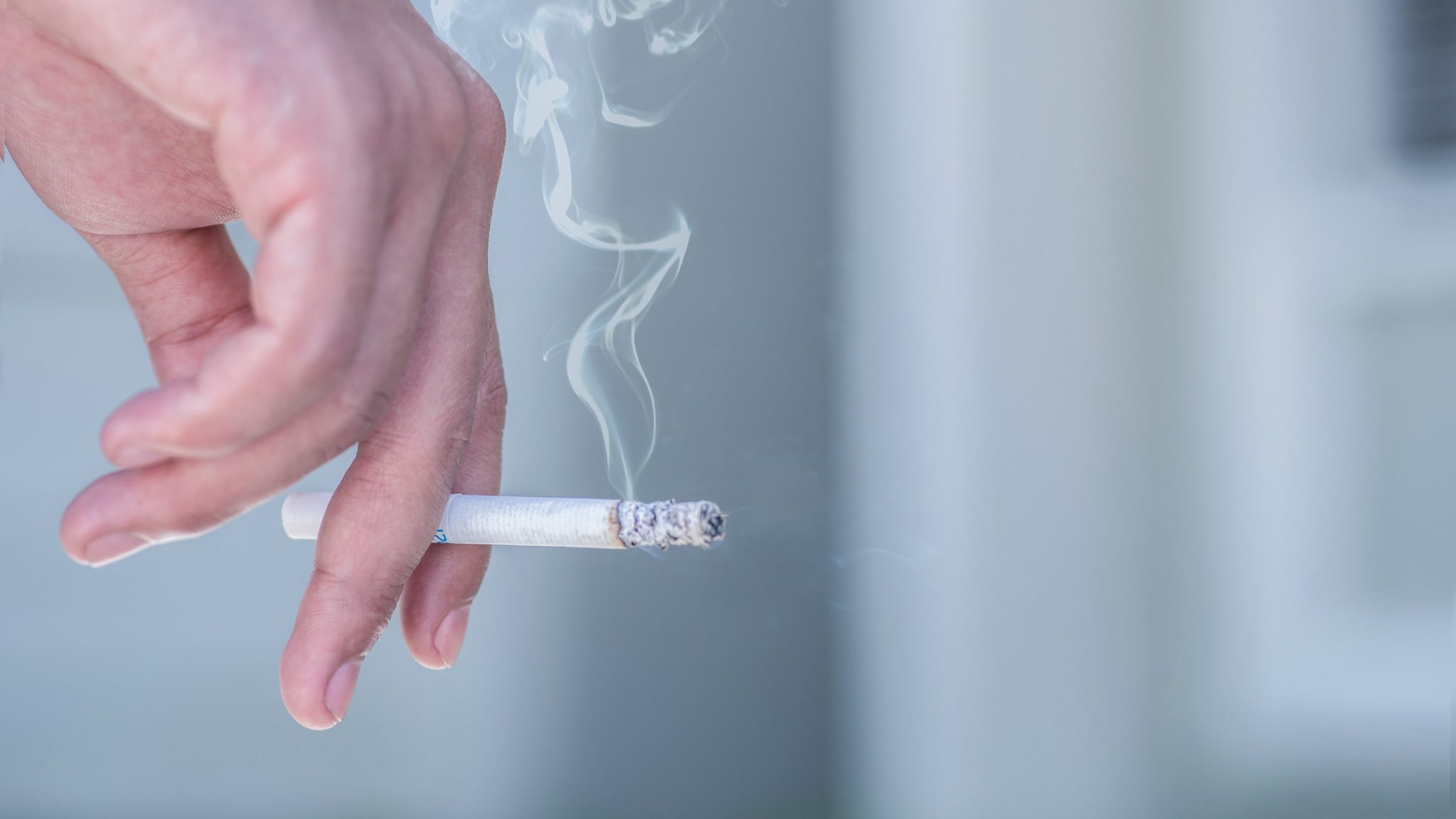 A hand holding a lit cigarette with cigarette smoke flowing upward.