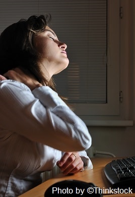 A woman sits at a desk with her hand on her sore neck