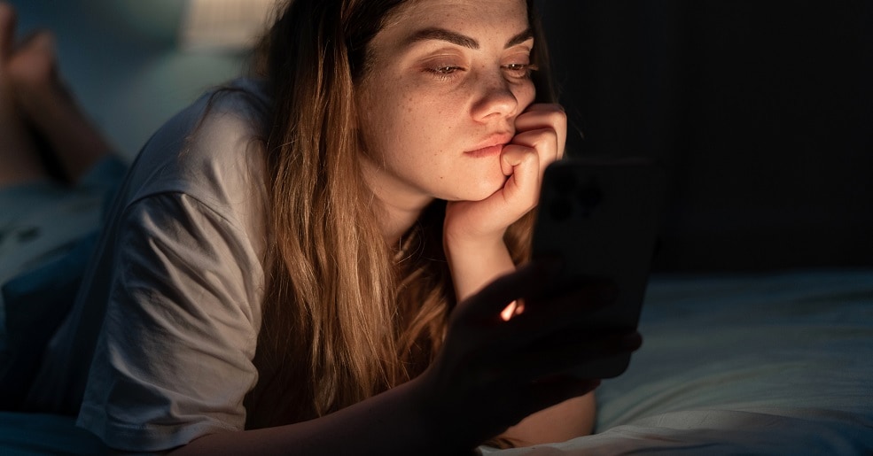 Woman sitting in dark in bed, looking at cell phone.