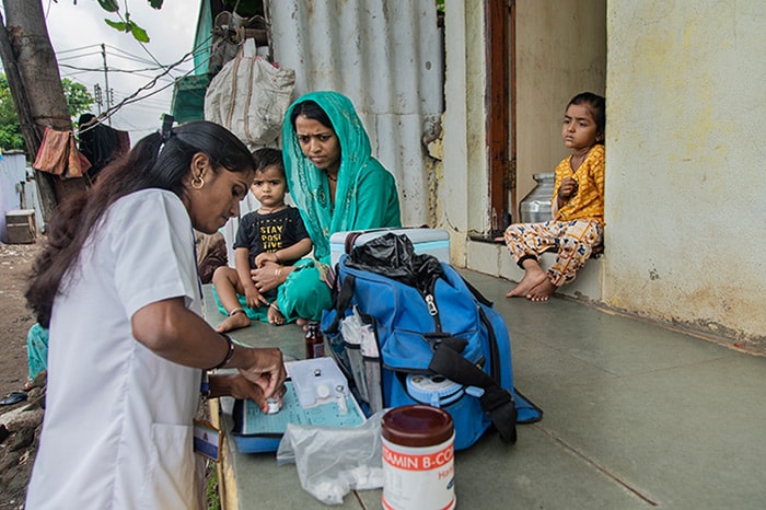 A health worker prepares medical supplies outside a home in India, watched by a mom and her children.