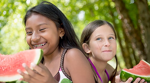 American Indian girl and White girl each smiling and eating a large slice of watermelon.