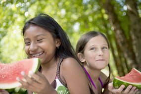 two pre-teen girls smiling and eating watermelon outside on a sunny day
